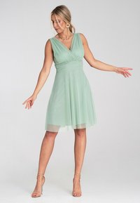 Mintgrünes ärmelloses Kleid mit Wickeloberteil, definiertem Taillenbereich und mehrlagigem Rock, aus weichem, fließendem Stoff und mit knieumspielendem Saum.