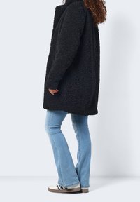 Svart fluffig kappa i texturerat tyg, med lös passform och hög krage, tillsammans med ljusblå utsvängda jeans och avslappnade sneakers.