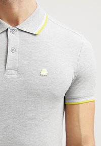 Grå pikétröja i texturerat material, med gula och vita detaljer på kragen och ärmarna, samt en liten broderad logotyp på bröstet.