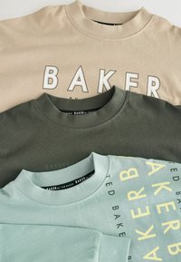 Drei gestapelte T-Shirts. Farben: beige, olivgrün, hellblau. Merkmale: runde Ausschnitte, weicher Baumwollstoff und "BAKER"-Druck in verschiedenen Stilen.
