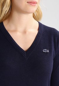 Kobieta w granatowym swetrze z dekoltem w serek, z małym haftowanym logo krokodyla na piersi, pokazana od ramion do podbródka.