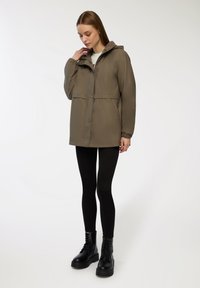 Giacca impermeabile khaki con cappuccio, polsini elasticizzati e chiusura frontale con bottoni. Abbinata a leggings neri e stivaletti da combattimento neri e robusti.