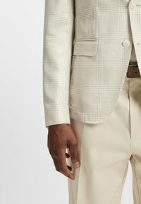 Beige Karo-Blazer aus strukturiertem Stoff, mit einem Reverskragen, zwei Vordertaschen und einem einzigen Knopfverschluss, kombiniert mit beigen Hosen.