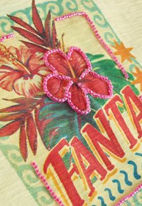 Fleur tropicale rouge et orange avec des feuilles vertes sur tissu, soulignée par des perles roses au-dessus du texte rouge en gras « FANAI » et une bordure décorative vert-bleu.