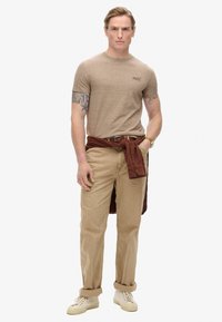 Camisa corta de manga con textura marrón, pantalones de pana beige y una camisa de color óxido atada a la cintura. Los zapatos son de un color claro y con un diseño sencillo.