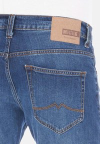 Jeans de mezclilla azul oscuro con una etiqueta de cuero marrón que dice "MUSTANG True Denim", costuras en contraste y un bolsillo trasero con un diseño de costura curvada.