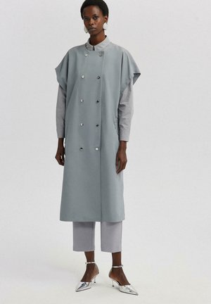 Manteau long à double boutonnage en tissu gris clair avec un col haut, doté de grandes boutons en argent et de poches latérales, associé à un pantalon court gris et des chaussures à talons métalliques.