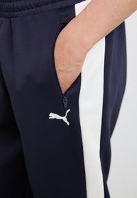 Marine joggebukser med en hvit side stripe, glidelåslomme og en liten hvit Puma-logo på låret. Laget av strukturt stoff.