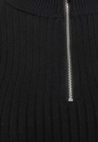 Haut en maille côtelée noire avec un col montant et un détail de fermeture éclair argentée au centre devant. Tissu texturé à lignes verticales.