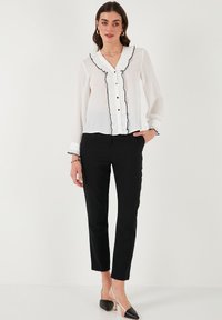 White blouse with black ruffle trim, V-neck, and buttons; paired with black tailored pants. Shoes are black and nude color-blocked with a heel.