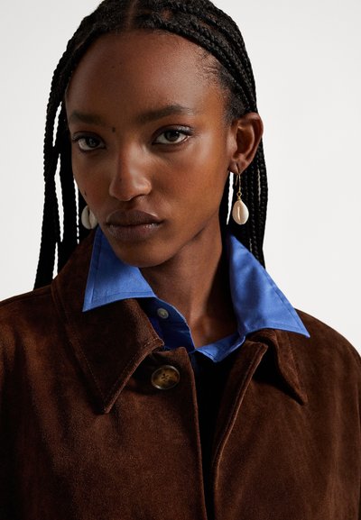 Hnedá semišová bunda s golierom a gombíkovými detailmi, vrstvená cez modrú košeľu. Dlhé zapletené vlasy, s bielymi náušnicami v tvare mušlí.