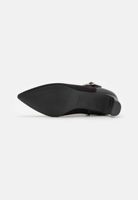 Zapato negro de ante de punta con suela lisa, tacón texturizado y un pequeño acento de hebilla metálica en el lateral.