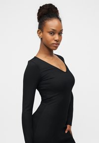 Μακρυμάνικο, μαύρο ριμπ φόρεμα με V-λαιμόκοψη, εφαρμοστή σιλουέτα και λεία υφή. Απλός σχεδιασμός που τονίζει την κομψότητα και την ευελιξία.