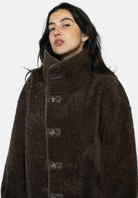 Bruine fleece jas met hoge kraag, voorzien van een glad leren bies en grote ronde knopen. Zachte textuur, oversized pasvorm met zichtbare naden.