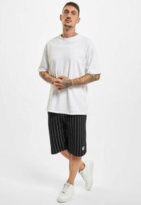 Tricou alb din bumbac cu mâneci scurte, asortat cu pantaloni scurți negri cu dungi și un logo alb. Stilizat cu adidași albi.