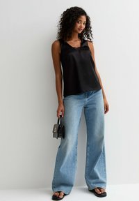 Zwarte satijnen mouwloze top met een v-hals en kanten details, gecombineerd met lichtblauwe wijde jeans en zwarte platte sandalen. Kleine zwarte handtas.