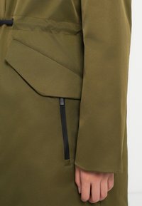 Cappotto verde oliva con una texture liscia, una grande tasca inclinata e una tasca con zip con hardware nero sul lato.