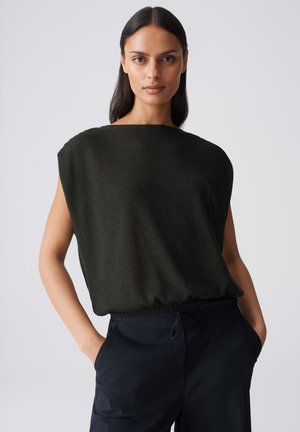 Donna con capelli lisci e scuri che indossa una canotta nera senza maniche e pantaloni scuri, in piedi con le mani in tasca su uno sfondo semplice.