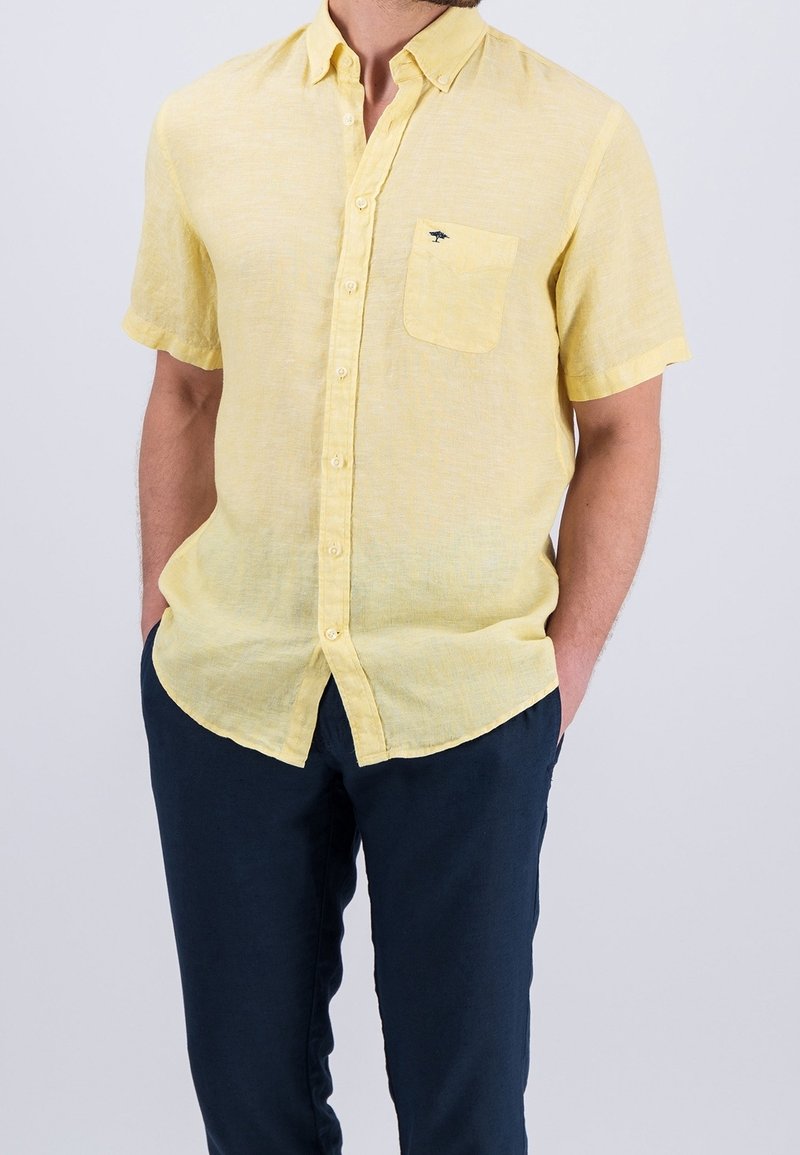 Chemise en lin jaune à manches courtes, avec une poche avant et une fermeture à boutons, associée à un pantalon bleu marine foncé. Tissu léger et texturé.