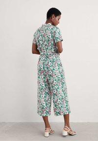 Ziedu kombinezons ar īsām piedurknēm, ar zaļu fona krāsu un rozā, zilu un baltu rakstu. Izgriezti gurni un josta ap vidukli.