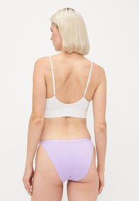 Mulher com cabelo loiro curto, vestindo um sutiã branco e uma roupa interior em estilo biquíni lilás, de pé contra um fundo branco liso.