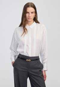 Weiß gestreiftes Hemd mit Knopfleiste und Kragen, kombiniert mit grauen maßgeschneiderten Hosen und einem schwarzen Gürtel mit goldener Kettenakzent.