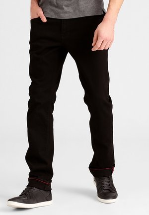 Homme portant un jean noir, une chemise grise et des baskets noires, debout devant un fond clair uni.