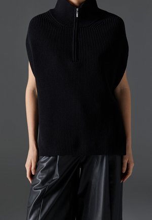 Personne portant un gilet en maille côtelée noir sans manches avec une fermeture éclair à l'avant et un pantalon ample en cuir noir sur fond sombre.