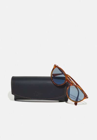 Occhiali da sole rotondi con montatura in tartaruga ambra e lenti blu, poggiati su un astuccio in pelle nera con cuciture a contrasto.