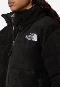 Czarna polarowa kurtka z wysokim kołnierzem i pełnym zamkiem, z białym haftowanym logo po lewej stronie klatki piersiowej oraz teksturowanym materiałem.