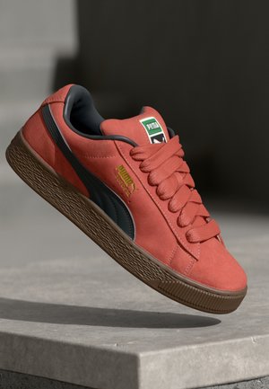 Baskets en daim corail avec accents noirs, bout rond, épaisse semelle en caoutchouc marron et lacets plats. Marque Puma sur la languette et le panneau latéral.