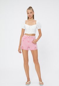 Top blanco cortado con mangas abullonadas y lazo en el frente. Pantalones cortos de mezclilla rosa desgastados con dobladillo deshilachado. Llevado con chanclas blancas.