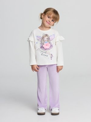 Giovane ragazza con capelli biondi che indossa un maglione bianco con una fata e la scritta "Sogni magici", pantaloni lavanda e scarpe bianche, che sorride leggermente.