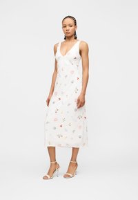 Γυναίκα που φοράει λευκό αμάνικο μίντι φόρεμα με βαθύ V λαιμόκοψη και πολύχρωμο σχέδιο με λουλούδια και κοχύλια, συνδυασμένο με μπεζ πέδιλα με ψηλό τακούνι.