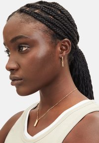 Guldtonede nøgleformede øreringe og en rektangulær vedhængs kæde bæres med en lys beige ærmeløs top, der viser struktureret stof.