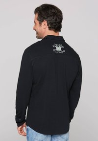 Camisa negra de manga larga con un acabado texturizado, que presenta un estampado gráfico en la parte superior de la espalda. Puños abotonados y diseño de dobladillo curvado.