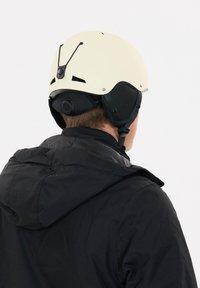 Hvid hjelm med mat finish, sorte ørepolstringer, justerbar rem og ventilationshuller, båret af en person i en sort jakke.