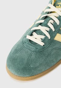 Zielone zamszowe sneakersy z żółtymi paskami, z białymi detalami na sznurówki i gumową podeszwą, prezentujące fakturowany materiał i przeszywane akcenty.