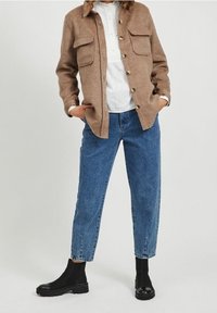 Brun, skjortejakke med knapper og to brystlommer over en hvid bluse, kombineret med blå cropped jeans og sorte ankelstøvler.