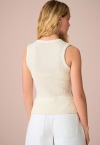 Top senza maniche in maglia color crema con texture a coste, vita aderente e scollo rotondo, abbinato a pantaloni bianchi a vita alta.