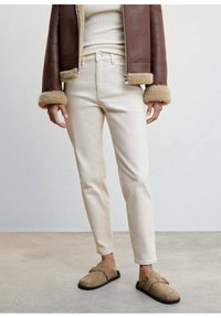 Jeans ajustados beiges con una cintura de media altura, combinados con una chaqueta marrón forrada de piel de oveja. Zapatos marrones de tipo slip-on completan el conjunto.