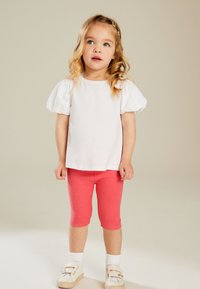 Witte katoenen top met pofmouwen, gecombineerd met koraalroze legging en crèmekleurige schoenen met klittenbandsluitingen en regenboogaccenten.
