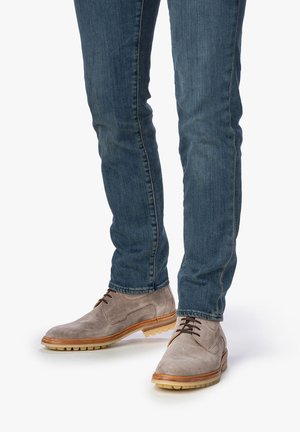 Zapatos de ante gris con detalles en cuero marrón, con punta redondeada y cordones. Llevados con jeans de mezclilla azul, que muestran un ajuste ceñido.