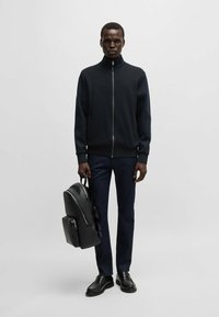 Black zip-up sweatshirt with high collar, paired with dark jeans and black leather shoes, holding a textured black backpack.