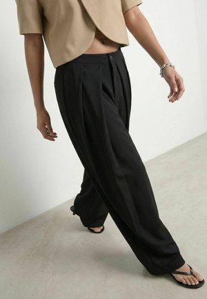 Persoon loopt met losse zwarte broek, beige cropped top en zwarte teenslippers, met een zilveren armband aan één pols.