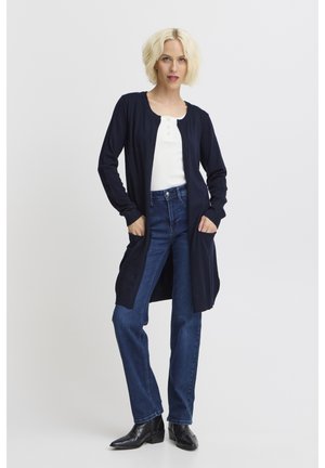 Mujer con cabello corto y rubio que lleva una camiseta blanca, un cárdigan largo azul marino, vaqueros azul oscuro y botines negros, de pie con las manos en los bolsillos.