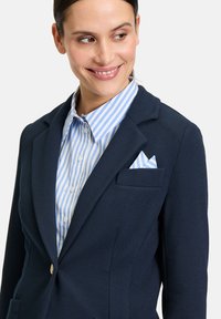 Granatowa marynarka o teksturowanej tkaninie, dopasowanym kroju i z pojedynczym zapięciem na guzik. W zestawie z niebieską koszulą w biało-niebieskie paski oraz poszetką.