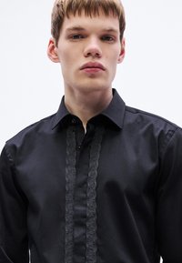 Fekete ing hagyományos gallérral, elöl texturált fekete szalagdíszítéssel. Sima anyagból készült, testhez simuló szabással.