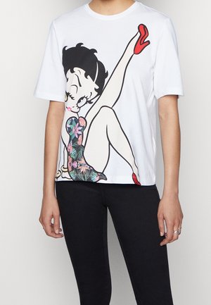 Biała koszulka z dużą kreskówkową kobietą o czarnych włosach, w kwiecistej sukience, czerwonych szpilkach, z jedną uniesioną nogą, noszona z czarnymi spodniami.