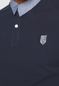 Muž v námořnickém modrém polo tričku s bílým knoflíkem a bílou vyšitou emblémem na hrudi, s světle modrým límečkem.